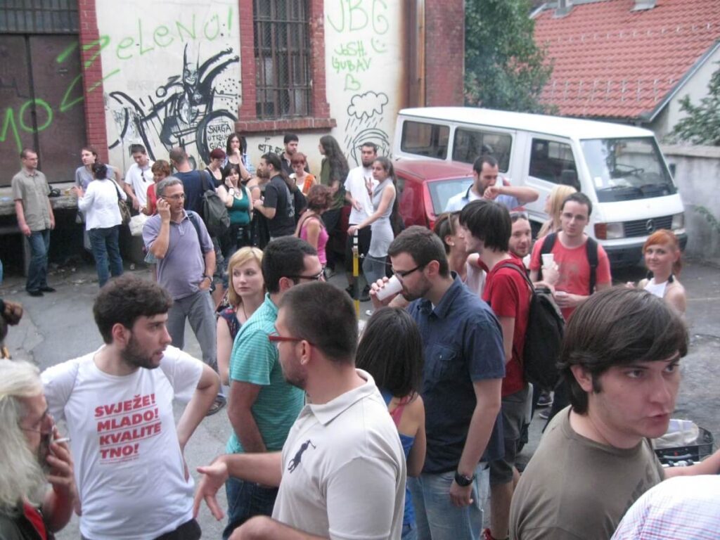 Велика група људи окупља се напољу близу зграде прекривене графитима, што одражава дух уметности и солидарности. Неки разговарају док други разгледају; аутомобили и бели комби су паркирани у близини, стварајући опуштену и друштвену атмосферу Контекст колектива.