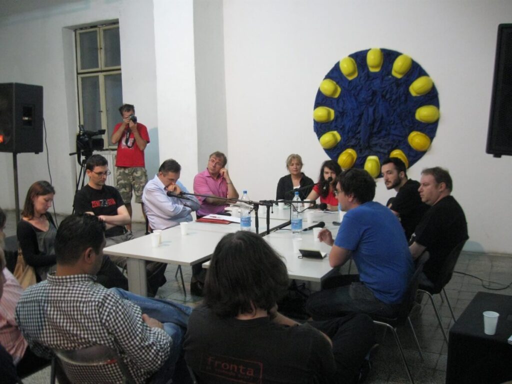 Група људи седи око белог стола на панелу за дискусију о уметности и активизму; неки говоре док други слушају. Иза њих, жуте кациге формирају круг на плавој позадини, док сниматељ снима овај тренутак колективне солидарности.