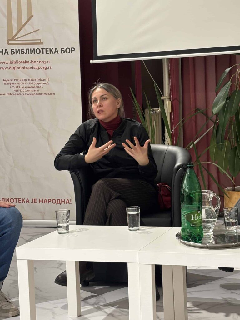 Жена седи у црној фотељи и гестикулира рукама док говори. Налази се унутра, поред белог стола са чашама, зеленом флашом и бокалом. Иза ње је транспарент и бордо завеса.