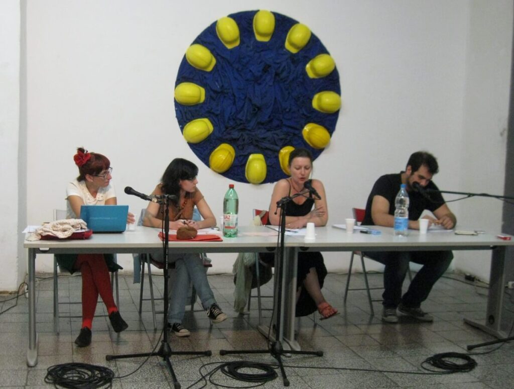 Четири особе седе за столом са микрофонима, говоре или читају. Иза њих, плави круг са жутим заштитним кацигама – симбол солидарности и активизма – краси зид. Флаше и папири су разбацани по столу.