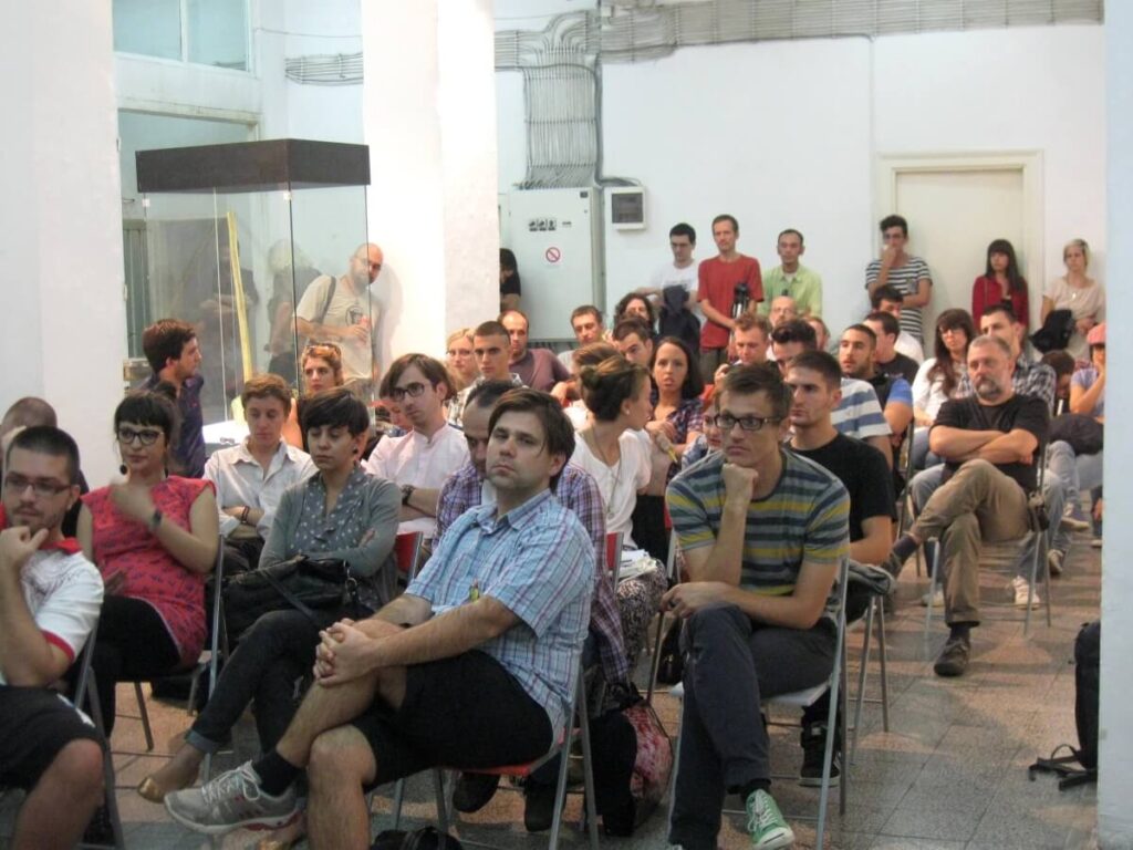 Група људи пажљиво седи у редовима столица у јарко осветљеној просторији, слушајући презентацију о уметности и активизму. Неки појединци се радују, док су други заокупљени размишљањем.