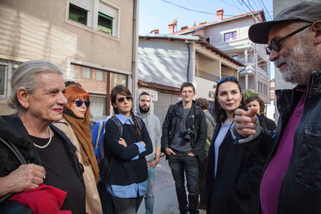 Група људи из Контекст колектива стоји и разговара на сунчаној улици са зградама у позадини. Један човек говори о уметности и активизму док други пажљиво слушају - неки са прекрштеним рукама, један држи камеру.