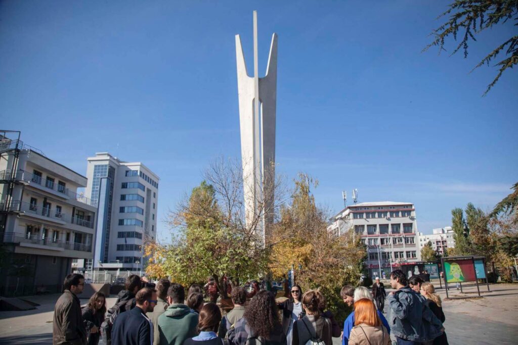 Група људи окупља се поред модерног, високог бетонског споменика окруженог дрвећем и зградама по сунчаном дану, отелотворујући уметност и активизам док стварају простор за колективну рефлексију под ведрим плавим небом.