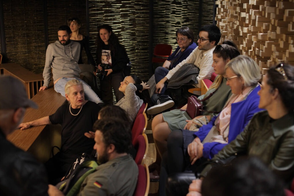 Група људи седи близу једна другој у просторији са дрвеним зидовима, заокупљена разговором или слушањем. Атмосфера одражава колективну солидарност, јер неки дискутују о уметности или активизму, док други пажљиво посматрају или комуницирају.