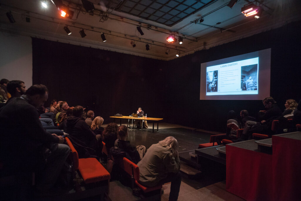 Група људи седи у слабо осветљеној просторији на црвеним столицама, посматрајући презентера за столом и великим екраном - догађај Контекст колектива који истражује уметност, солидарност и активизам кроз занимљиве презентације.