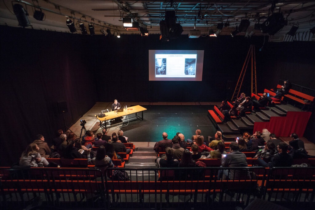 Особа седи за столом на бини и држи презентацију о уметности и активизму публици која седи на клупама распоређеним на више нивоа. Велики екран иза презентера приказује слајдове у мрачној просторији, налик биоскопској.