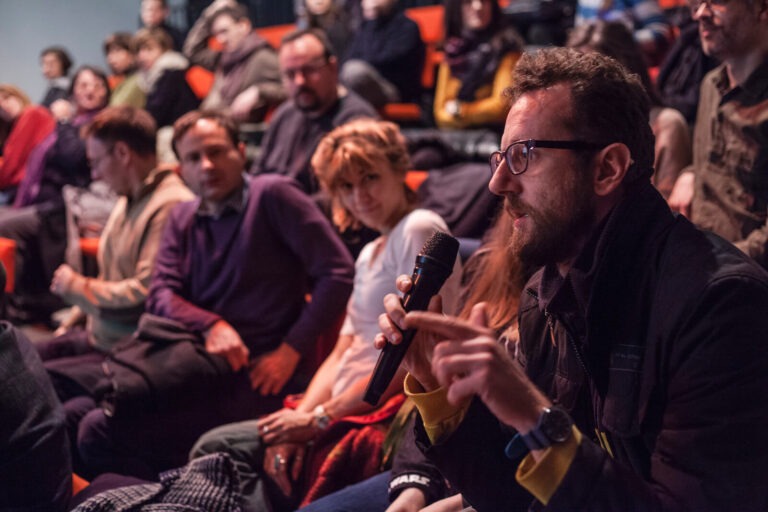 Човек са наочарима држи микрофон и говори током дискусије о активизму у седећој публици. Остали присутни, неки гледају у њега, а други радосно гледају унапред, испуњавају позадину просторије.