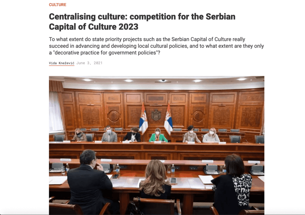 Група људи седи за великим дрвеним конференцијским столом у формалној сали за састанке са српским заставама и плавом заставом иза њих, разговарајући о уметности и активизму. Зидови обложени дрветом и микрофони истичу званични контекст њиховог окупљања.