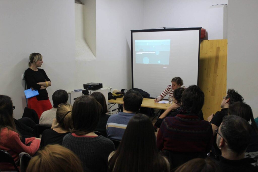 Група људи седи окренута ка жени која држи презентацију поред особе која седи за столом, са пројектованим платном на којем се приказује презентација о уметности и солидарности у малој просторији са белим зидовима.