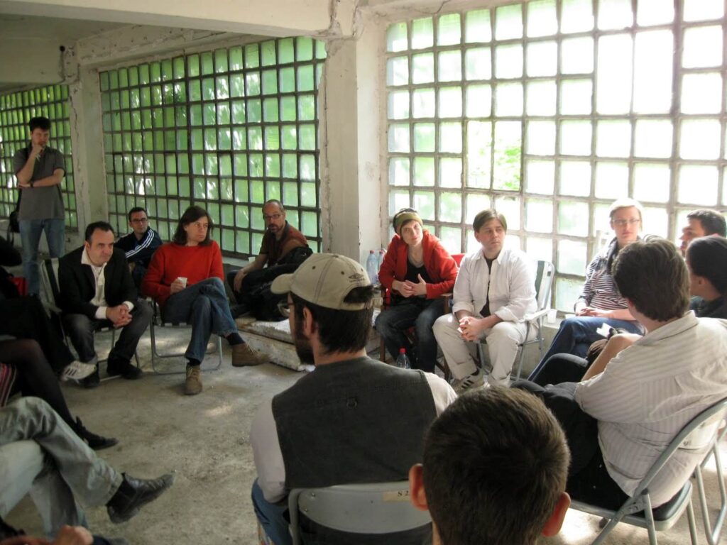 Група људи седи у кругу на склопивим столицама у светлој просторији индустријског стила са прозорима од стаклених блокова, учествујући у дискусији о уметности и солидарности.