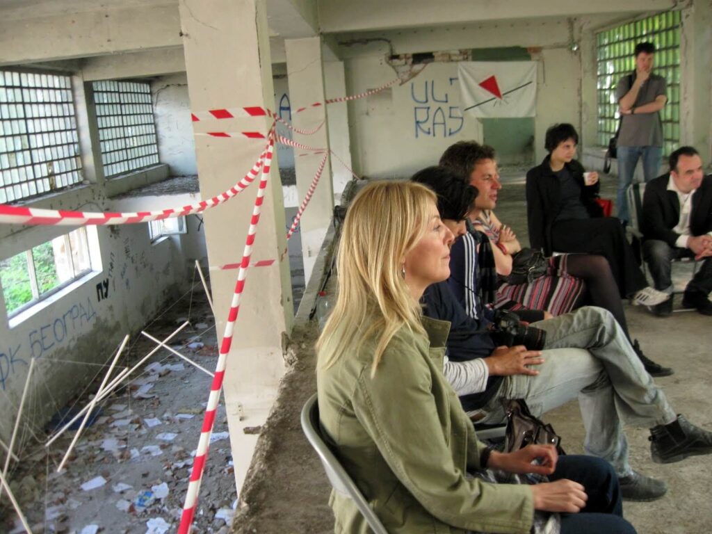 Група људи седи на столицама у трошној, графитима прекривеној просторији, подељеној црвено-белом траком за упозорење. Као део Контекст колектива, окупљају се због уметности и активизма, негујући солидарност усред напуштеног, претрпаног окружења.