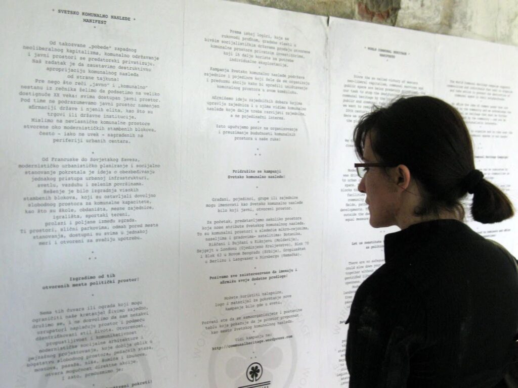 Особа са тамном косом и наочарима чита велики зидни дисплеј прекривен страницама штампаног текста на страном језику, истражујући теме уметности и солидарности. Дисплеј је постављен на белој позадини у добро осветљеном затвореном простору.