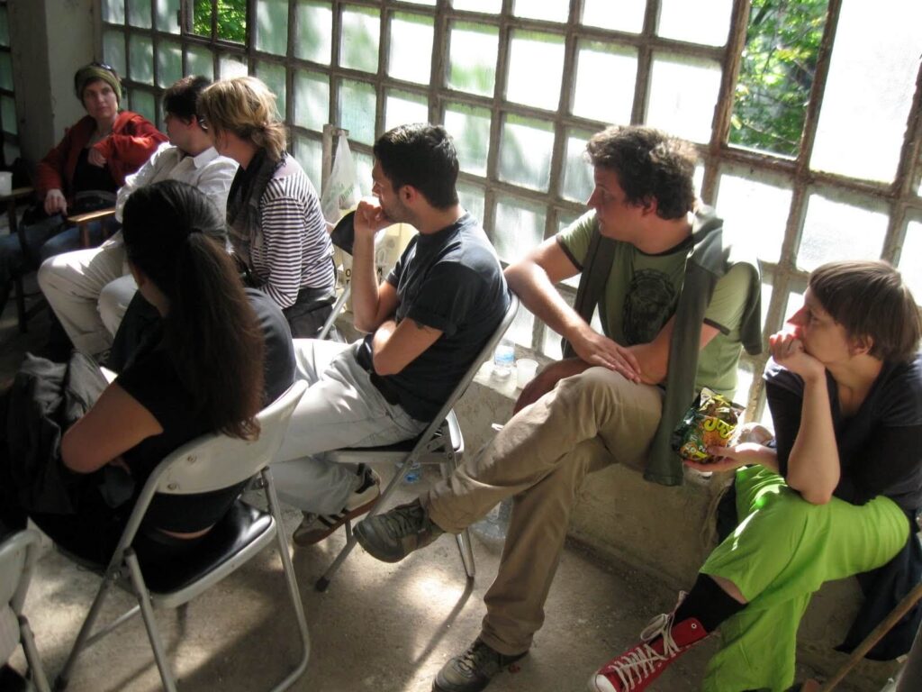 Група људи седи на белим склопивим столицама у сунцем обасјаној соби са прозорима од стаклених блокова, пажљиво слушајући некога ван видокруга. Неки имају свеске; једна особа једе грицкалице – отелотворујући уметност и солидарност кроз заједнички активизам.