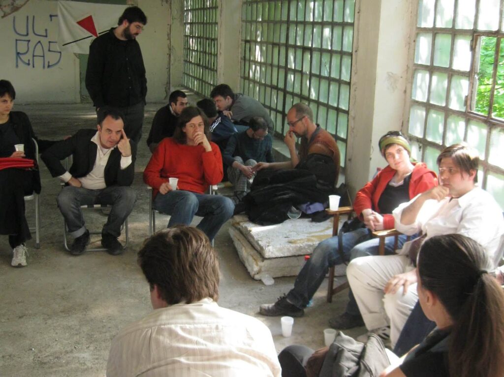 Група људи из Контекст колектива седи у сунцем обасјаној, недовршеној просторији са великим прозорима од стаклених блокова, учествујући у дискусијама о уметности и активизму. Графити исцртавају зид, а шоље леже на поду, стварајући атмосферу солидарности.