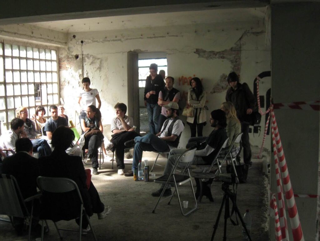 Група људи седи у кругу склопивих столица у истрошеној, индустријској просторији са изложеним циглама и залепљеним деловима, слушајући некога како говори; сунчева светлост струји кроз стаклене прозоре - атмосфера набијена уметношћу и солидарношћу.