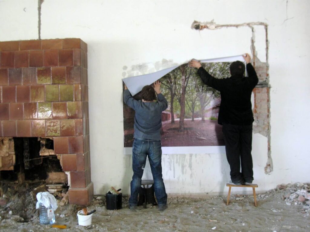 Двоје људи из Контекст колектива причвршћују велики постер стазе са дрворедом на оштећени зид у соби која се реновира, окружени алатима, грађевинским материјалом и знацима уметности и солидарности у свом заједничком активизму.