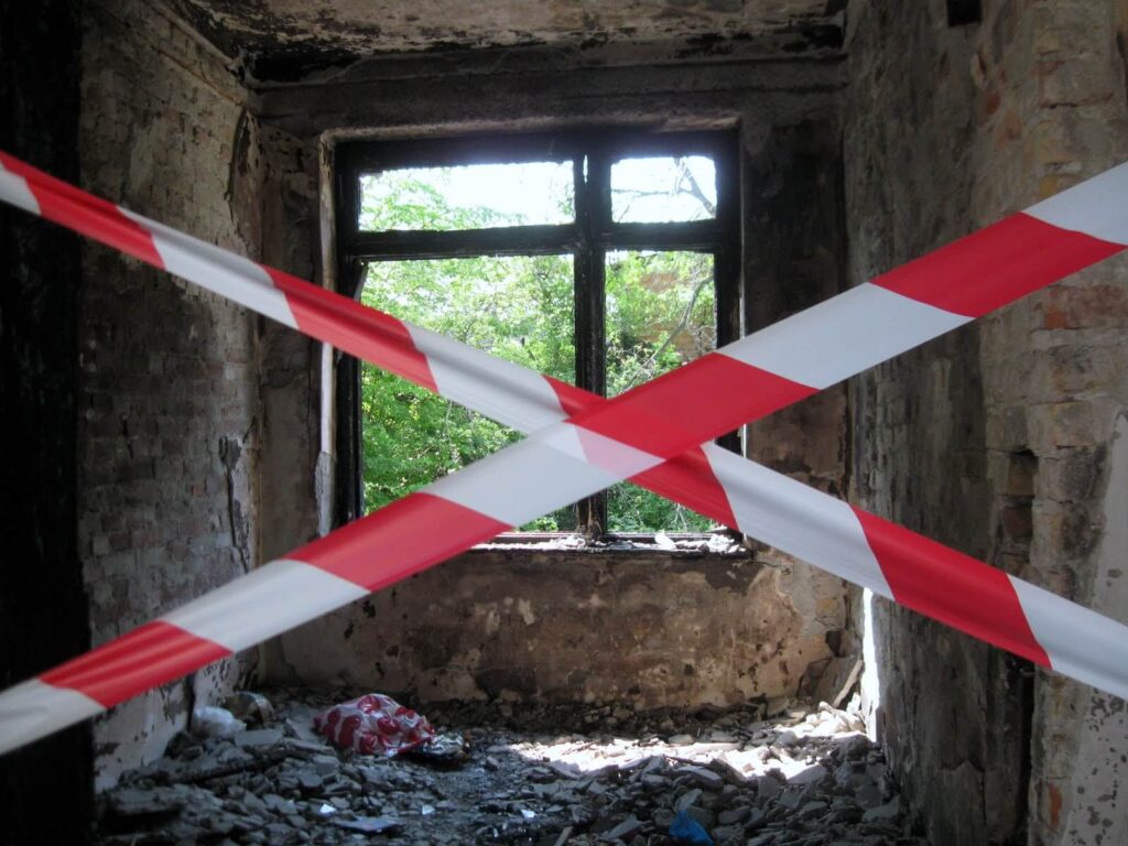 Трошна, напуштена соба са распадајућим зидовима и отпадом на поду, блокирана црвено-белом траком за упозорење — сунчева светлост и зеленило струје кроз разбијени прозор, наговештавајући уметност и солидарност оживљену активизмом.