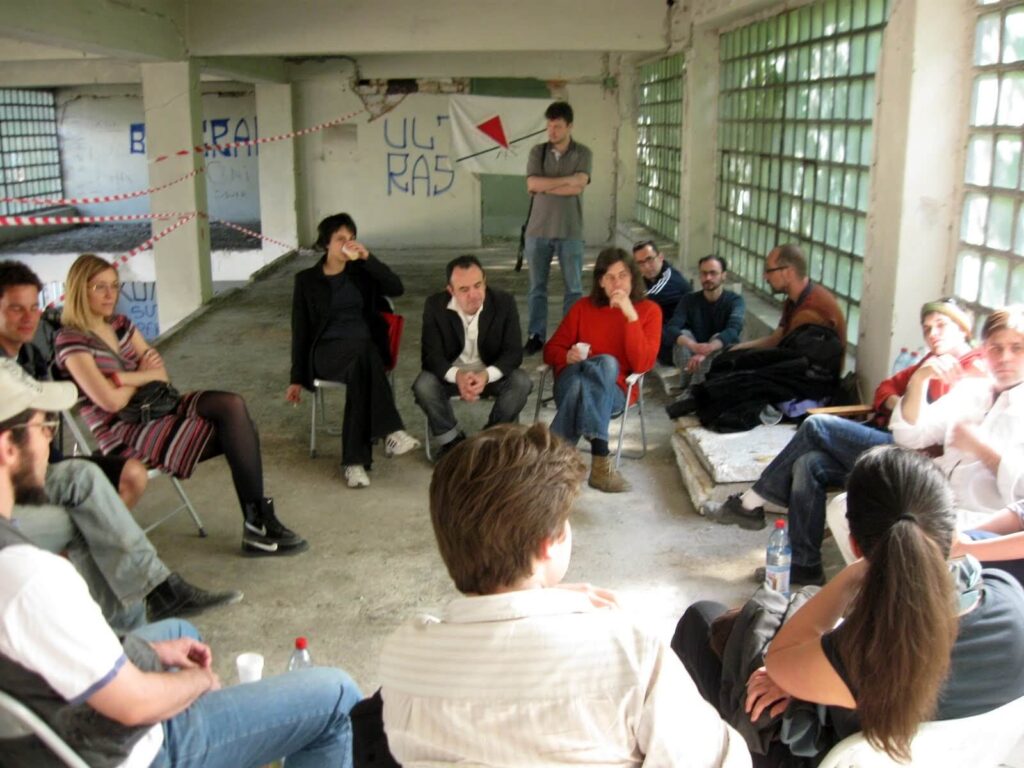 Група људи седи у кругу на столицама и клупама у великој, индустријски изгледајућој просторији са графитима на зидовима и великим прозорима, ангажовани у дискусији о уметности и солидарности унутар Контекст колектива.