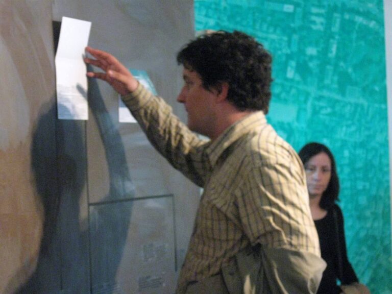 Мушкарац у карираној кошуљи интерагује са изложбом на зиду — вероватно део пројекта Контекст колектив уметност — држећи картицу, док жена стоји у позадини испред цијан мапе или графике.