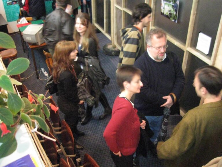 Група људи, од којих неки стоје и разговарају у паровима, интерагују у затвореном простору са тамним зидовима и урамљеним изложбама; инспирисани колективном уметношћу, они отелотворују солидарност док држе јакне, са биљком видљивом у првом плану.