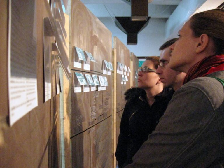 Троје људи из Контекст колектива пажљиво испитују паное са одштампаним документима и малим фотографијама, изгледајући као да проучавају или читају о уметности, солидарности и активизму у затвореном простору.