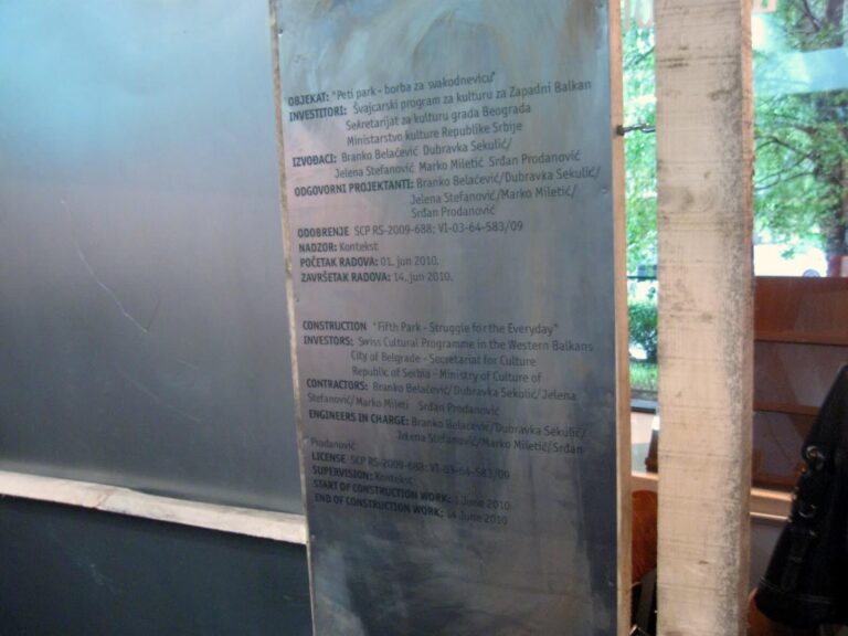 Метална плоча са угравираним двојезичним текстом на српском и енглеском језику пружа детаље о грађевинском пројекту. Креирао ју је Контекст колектив, а истиче уметност, солидарност и активизам. Дрвеће је видљиво у позадини.