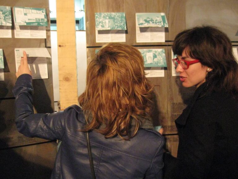 Две жене стоје унутра и гледају пано са фотографијама и натписима. Ангажоване уметношћу и активизмом, једна показује док друга, носећи црвене наочаре, слуша – што одражава дух солидарности који је кључан за Контекст колектив.