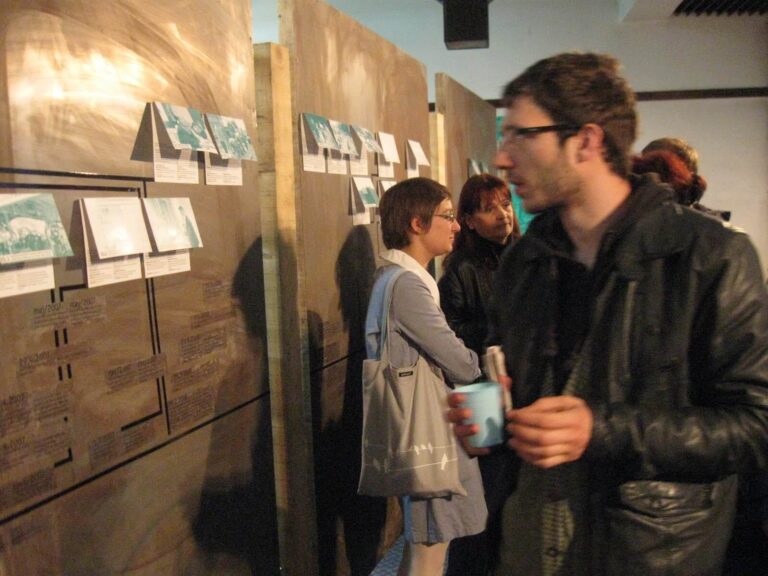 Људи посматрају уметничка дела и информативне панеле Контекст колектива, истичући уметност, солидарност и активизам. Једна особа у првом плану држи шољу док други пажљиво испитују експонате у овој лежерној, интерактивној изложби у затвореном простору.