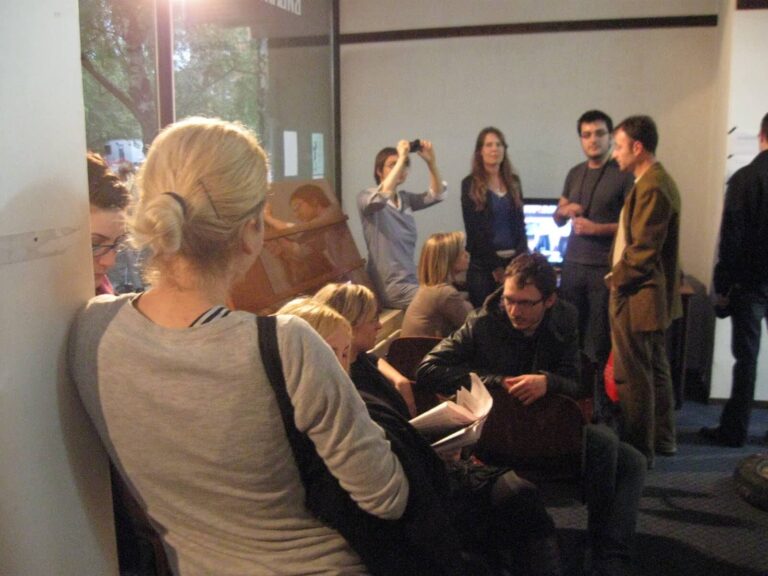 Група људи из Контекст колектива комуницира у сунцем обасјаној просторији са великим прозорима; неки стоје и разговарају о уметности и активизму, једна особа фотографише, док други седе, читају или ћаскају, стварајући атмосферу солидарности.