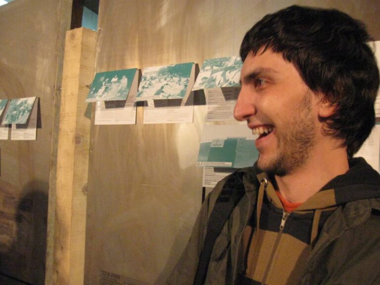 Човек са тамном косом и пругастом дуксерицом се осмехује док стоји унутра. Иза њега, неколико папира са сликама и текстом – који приказују уметност и активизам – изложени су на зиду под топлим, меким осветљењем.