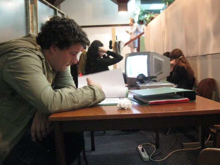 Човек у зеленој јакни седи за столом и чита књигу. Иза њега, две особе користе десктоп рачунаре, а трећа особа стоји на степеницама. Соба, обликована контекст колективом и активизмом, изгледа као канцеларија или простор за учење.