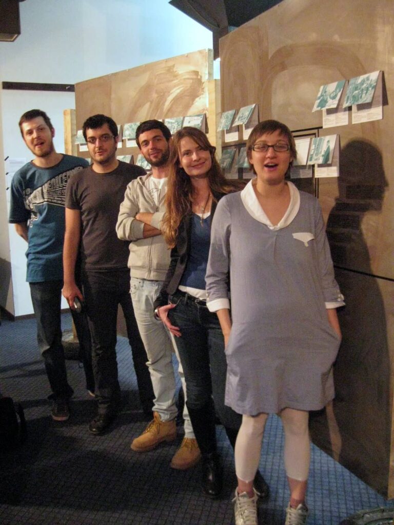 Петоро људи стоји унутра у реду, поређаних по степеништу, осмехујући се и лежерно позирајући. Уметничка дела су изложена иза њих, одражавајући посвећеност Контекст колектива уметности, солидарности и активизму у живахном галеријском окружењу.