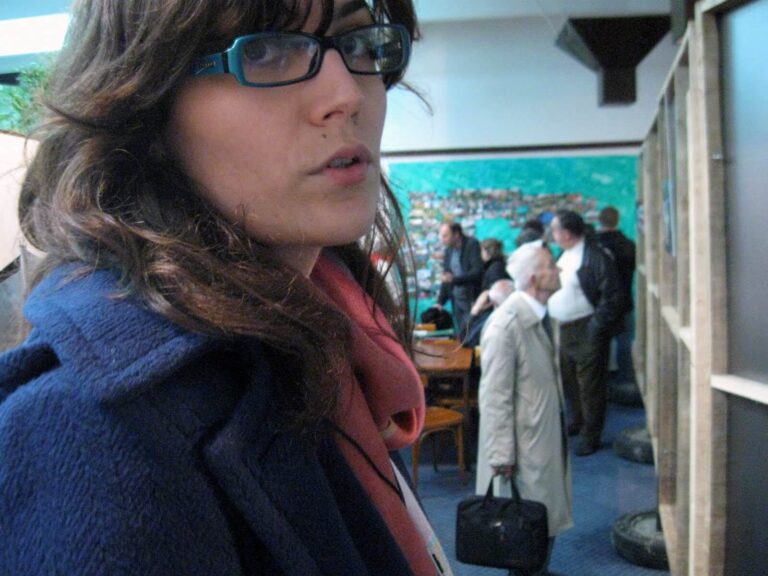 Жена са таласастом смеђом косом и наочарима гледа у камеру. Иза ње, чланови Контекст колектива стоје у соби са плавим зидовима и муралом, евоцирајући уметност и солидарност својим присуством и активизмом.