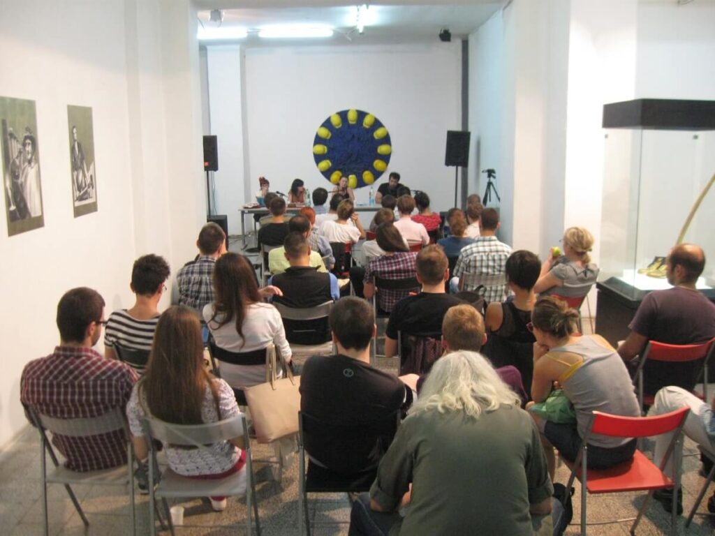 Чланови публике који седе у редовима окренути су ка панелу говорника у предњем делу просторије налик галерији, где Контекст колектив негује уметност и солидарност, окружен белим зидовима, изложеним уметничким делима и упечатљивим кружним уметничким делом на задњем зиду.