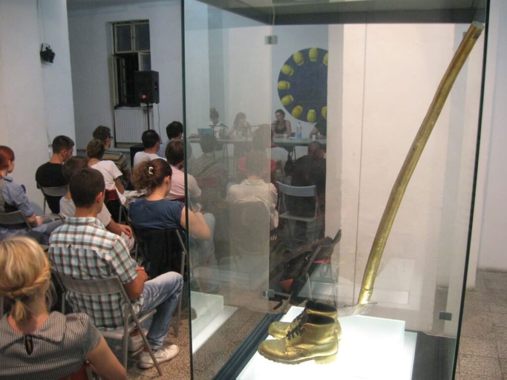 Публика слуша панел дискусију о уметности и активизму, са стакленом витрином у првом плану у којој се налазе велики златни штап и чизме. Плаво уметничко дело са жутим елементима виси иза звучника.