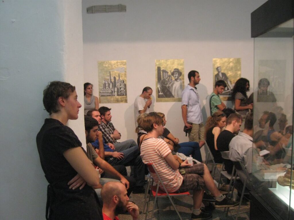 Група људи седи и стоји у малој галерији, пажљиво посматрајући нешто ван видокруга. Неки праве белешке, а уметничка дела Контекст колектива су изложена на зиду иза њих, истичући уметност и активизам.