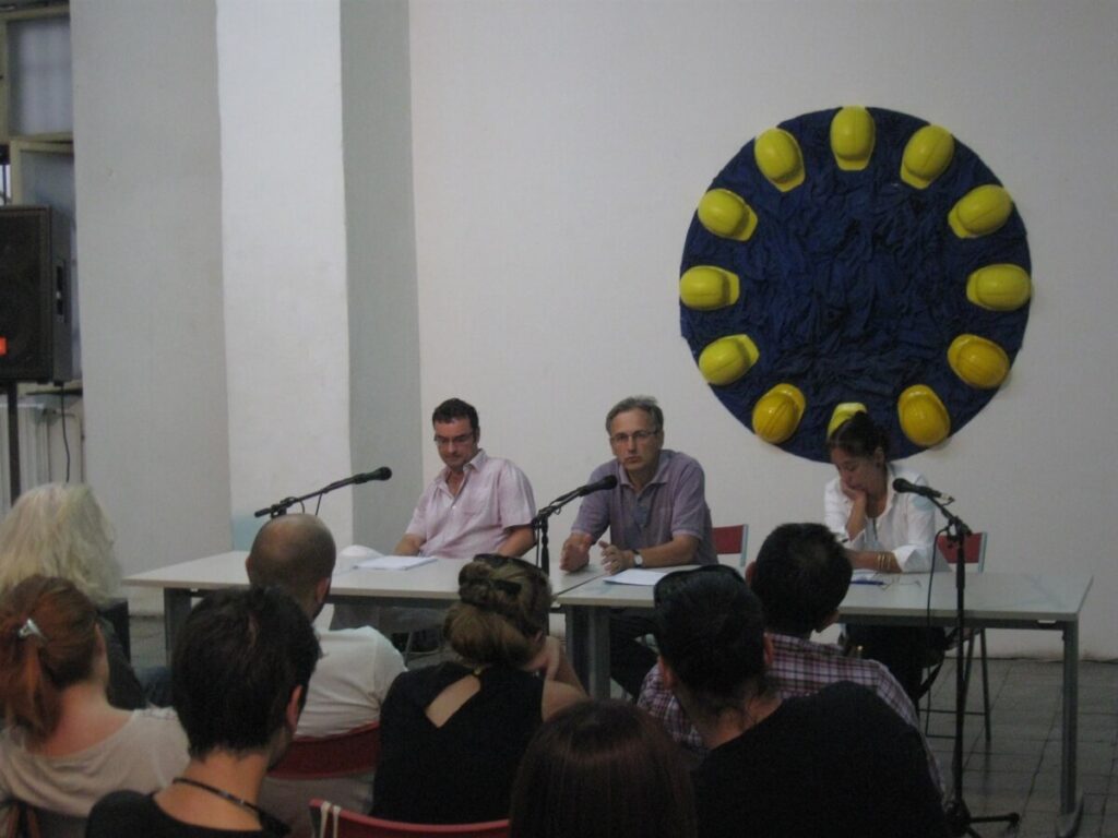 Троје људи из Контекст колектива седе за столом са микрофонима, ангажујући публику у дискусији о уметности, солидарности и активизму. Иза њих, жуте заштитне кациге формирају кружно уметничко дело насупрот тамноплавом зиду у минималистичкој просторији.