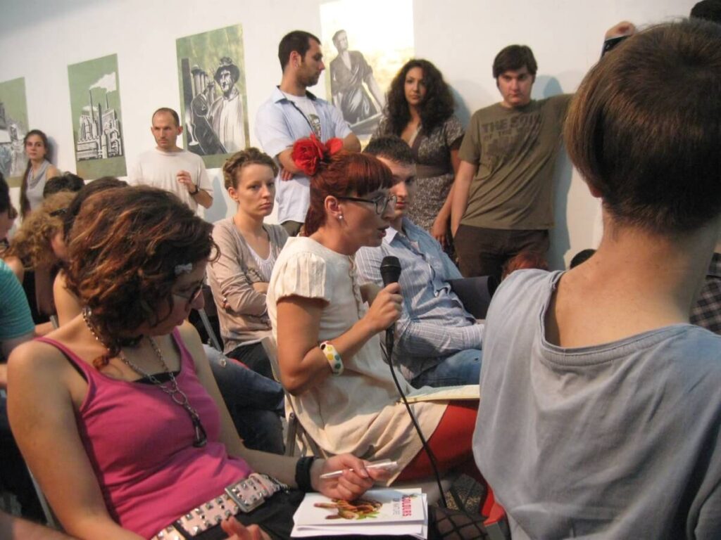 На догађају Контекст колектива, жена у белој хаљини са црвеним цвећем у коси држи микрофон, покрећући групу у дискусији о уметности и солидарности. Присутни пажљиво слушају, окружени уметничким делима на зиду.