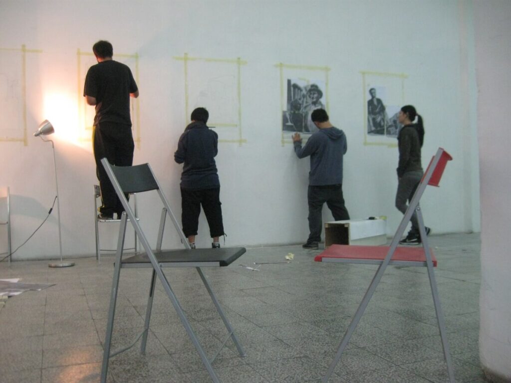 Четири особе из Контекст колектива окренуте су ка белом зиду, баве се уметношћу и активизмом док каче или цртају слике. Две столице, једна црна и једна црвена, седе у првом плану, док лампа са леве стране баца светло на ову сцену креативности и солидарности.