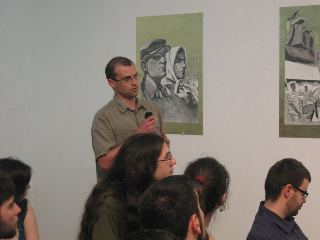 Човек који држи микрофон стоји међу људима који седе, говорећи — промовишући уметност и солидарност — испред два постера са монохроматским цртежима лица и фигура на белом зиду.