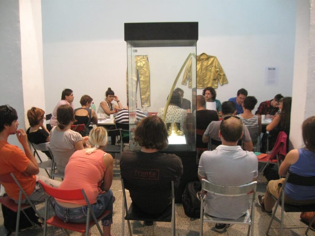 Група људи седи у кругу на склопивим столицама око стаклене витрине у јарко осветљеној просторији; две златне јакне су постављене на задњем зиду, истичући колективну уметност и дух солидарности.