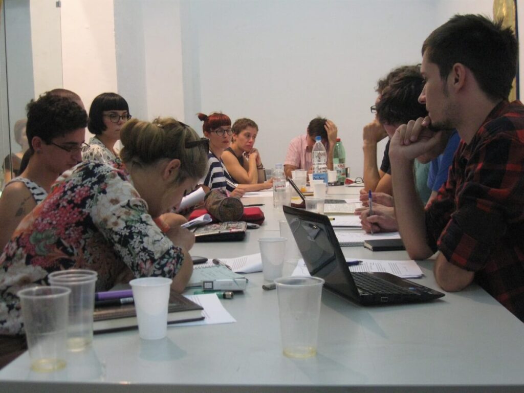 Група људи седи око дугачког стола прекривеног папирима, свескама, пластичним чашама и лаптоповима, ангажовани у колективној дискусији или проучавању о уметности и активизму у светлом затвореном окружењу.