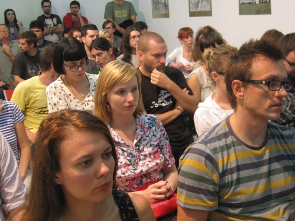 Група људи седи близу једни другима у јарко осветљеној просторији, пажљиво слушајући нешто ван кадра. Фокусирано расположење и ангажовани изрази лица сугеришу семинар или јавни говор о уметности или активизму у контексту колектива и солидарности.