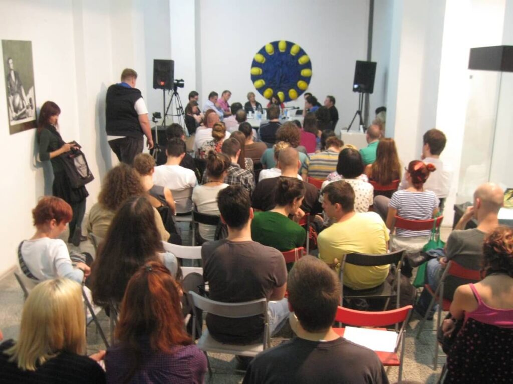 Група људи седи у редовима окренута је ка панелу говорника у предњем делу јарко осветљене просторије, са плаво-жутим кружним уметничким делом на зиду иза панелиста, које одражава уметност и солидарност на овом окупљању Контекст колектива.