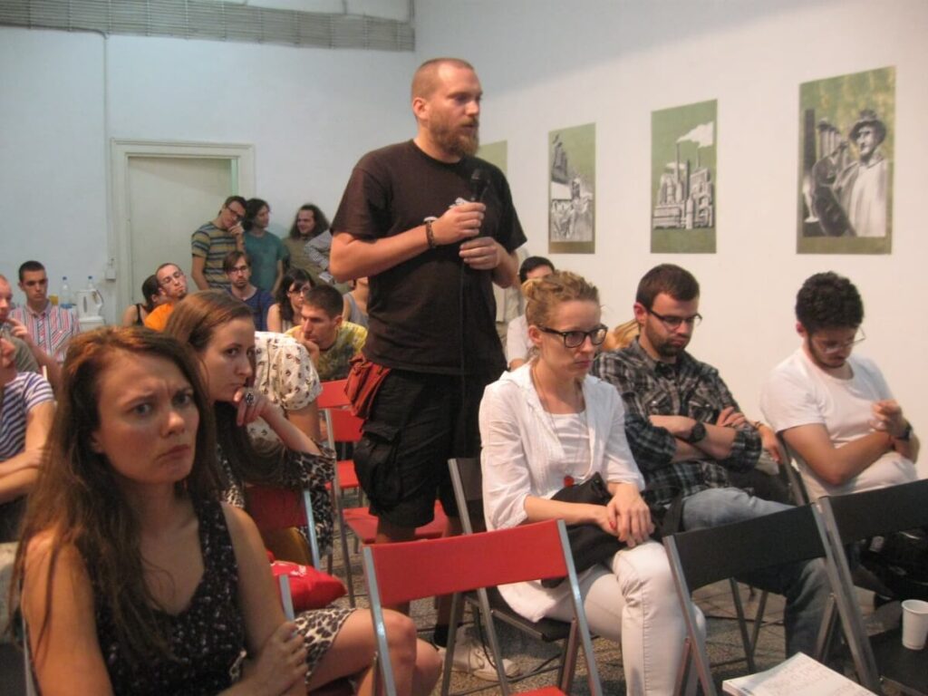 Човек стоји држећи микрофон, говорећи пред људима који седе - неки су пажљиви, други замишљени или расејани. Црно-бело уметничко дело Контекст колектива виси на белом зиду, одражавајући теме уметности и активизма.