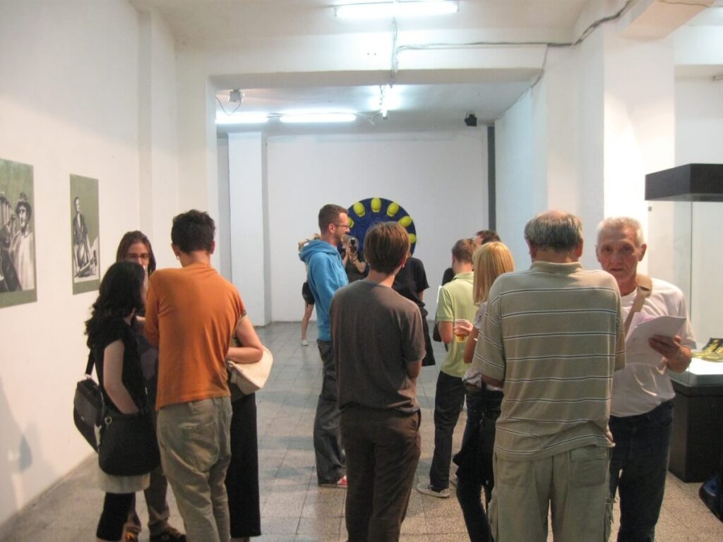 Група људи стоји и разговара у јарко осветљеној уметничкој галерији, дискутујући о уметности и активизму окружени сликама на зидовима и великим кружним уметничким делом на крају собе.