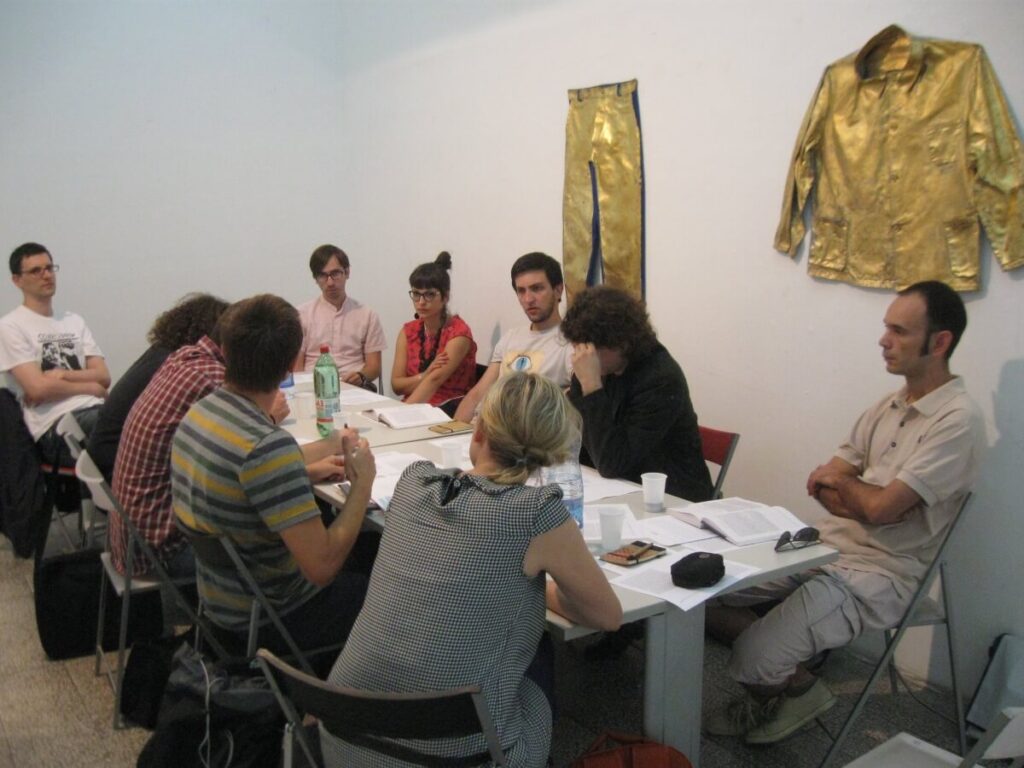 Група од девет људи седи око стола прекривеног папирима, оловкама и пићима, заокупљени дискусијом о уметности и солидарности. Два златна одевна предмета висе на белом зиду иза њих.