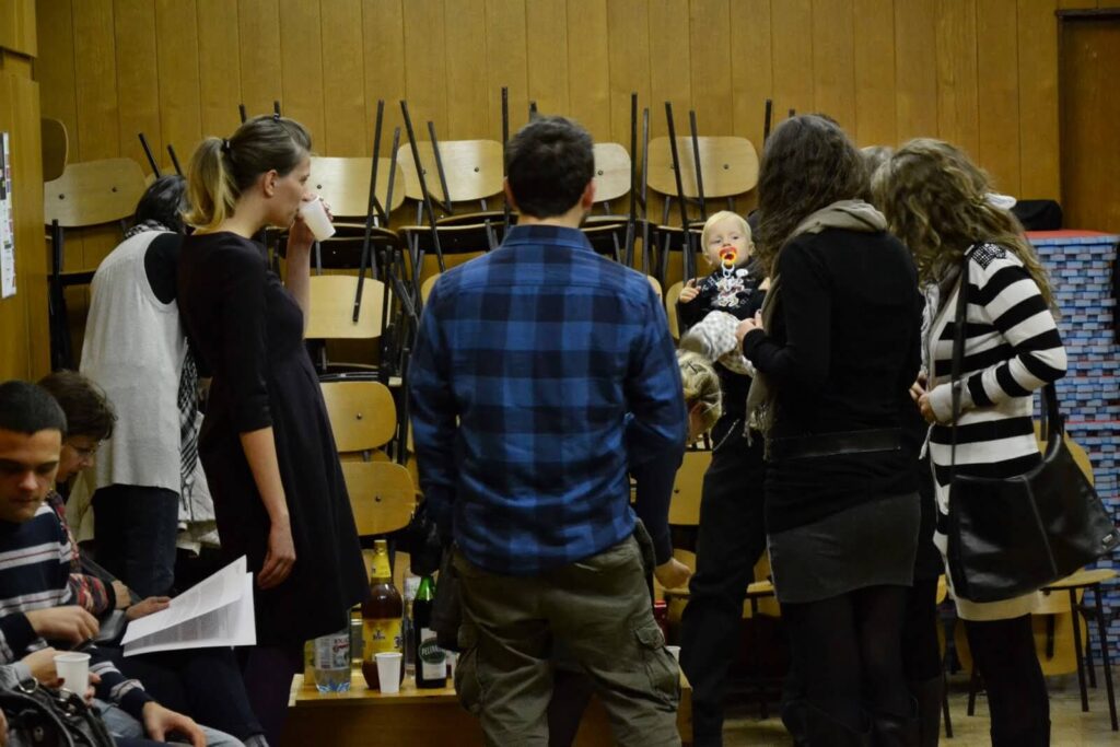 Група људи стоји у соби са дрвеним зидовима и наслаганим столицама; једна особа држи бебу са цуцлом, док други ћаскају и пију из шољица — стварајући топли контекст колектива, где се уметност, солидарност и активизам спајају.