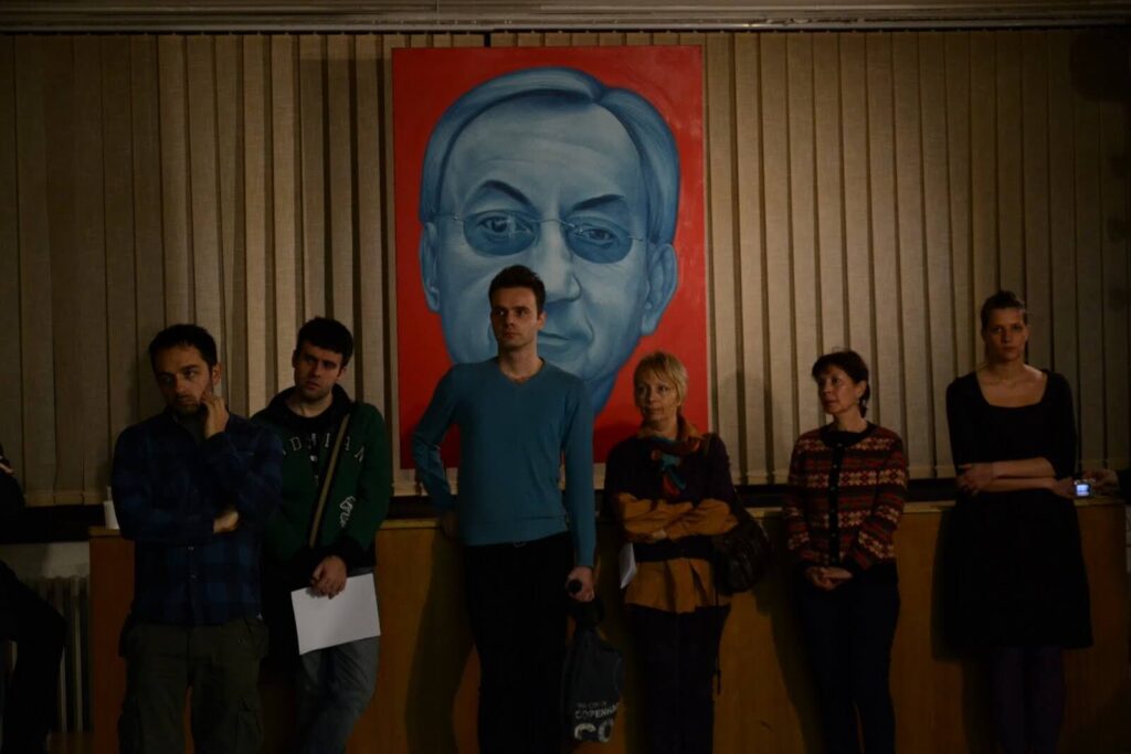 Шест људи стоји у слабо осветљеној просторији испод велике слике мушког лица на јаркоцрвеној позадини, са вертикалним жалузинама иза њих – евокативна сцена која одражава посвећеност Контекст колектива уметности и активизму.