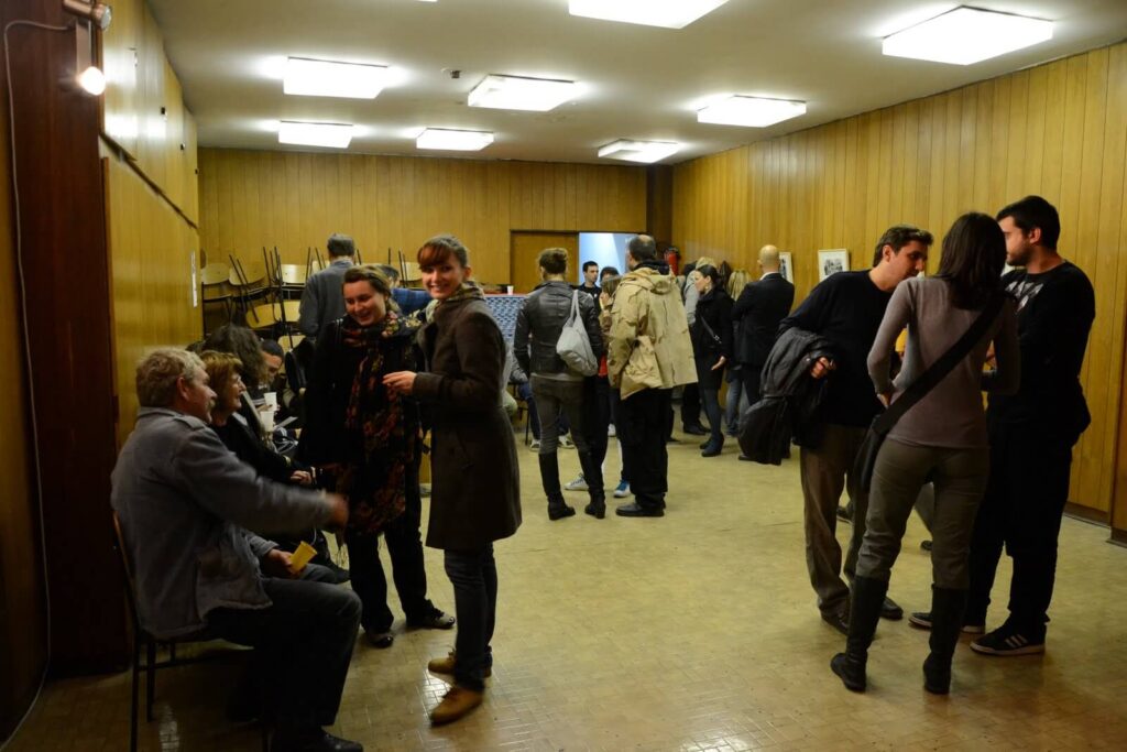 Група људи се дружи у једноставној просторији обложеној дрветом и са јарким плафонским светлима – окупљање које одражава вредности контекст колектива, где уметност и солидарност инспиришу разговоре међу учесницима који седе и стоје. Столице су наслагане једна на другу у позадини.